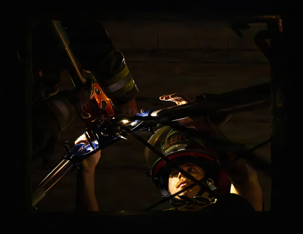 A dramatic, low-light image showing what appears to be a firefighter or rescue worker wearing a helmet with partial text visible, positioned in a confined or tight space. A second person in firefighting gear with reflective striping is visible above, and metallic rescue tools or equipment are prominently lit in the foreground. The scene suggests an active rescue operation or training exercise in a dark, enclosed environment.