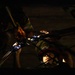 A dramatic, low-light image showing what appears to be a firefighter or rescue worker wearing a helmet with partial text visible, positioned in a confined or tight space. A second person in firefighting gear with reflective striping is visible above, and metallic rescue tools or equipment are prominently lit in the foreground. The scene suggests an active rescue operation or training exercise in a dark, enclosed environment.