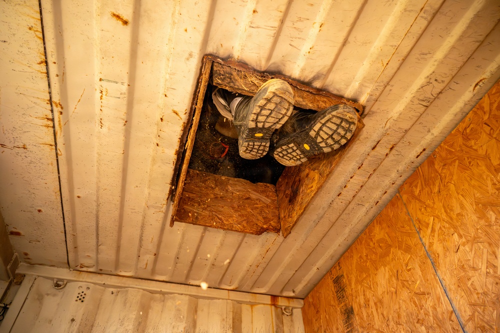 The image shows a person's legs and boots visible through a rectangular hatch or access opening cut into a ceiling. The boots have distinctive yellow dot markings on their soles and appear to be work or safety boots. The surrounding structure appears to be a metal-walled enclosure with OSB/plywood paneling on one side.