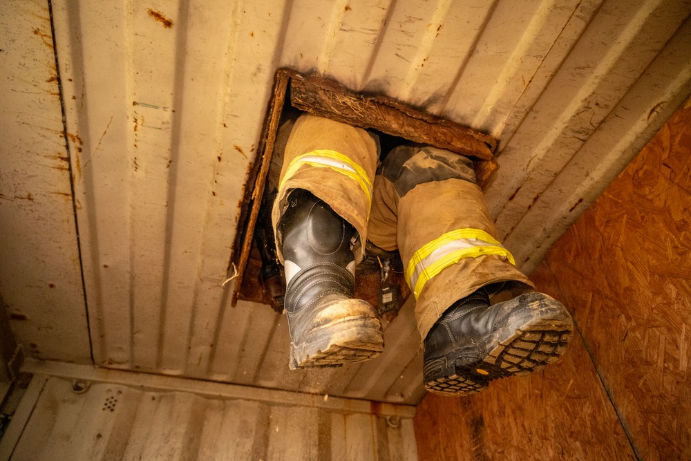 The image shows a low-angle view of a person's legs and boots descending through a hole cut in a ceiling or floor panel. The individual is wearing firefighter turnout gear, identifiable by the tan/brown protective pants with yellow reflective striping. The boots are heavily soiled black leather safety boots consistent with firefighting or emergency response equipment.