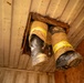 The image shows a low-angle view of a person's legs and boots descending through a hole cut in a ceiling or floor panel. The individual is wearing firefighter turnout gear, identifiable by the tan/brown protective pants with yellow reflective striping. The boots are heavily soiled black leather safety boots consistent with firefighting or emergency response equipment.