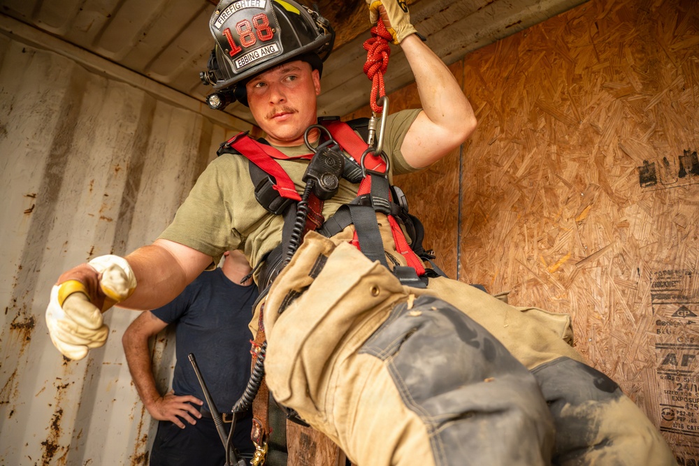 A firefighter wearing helmet number 188 from Ebbing ANG is engaged in a rope rescue or rappelling training exercise inside what appears to be a confined space or training structure. The firefighter is equipped with a red safety harness connected to a carabiner and rope system overhead, wearing standard turnout gear and protective gloves. The setting appears to be a training scenario conducted inside a plywood-lined metal container or mock structure.