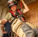 A firefighter wearing helmet number 188 from Ebbing ANG is engaged in a rope rescue or rappelling training exercise inside what appears to be a confined space or training structure. The firefighter is equipped with a red safety harness connected to a carabiner and rope system overhead, wearing standard turnout gear and protective gloves. The setting appears to be a training scenario conducted inside a plywood-lined metal container or mock structure.