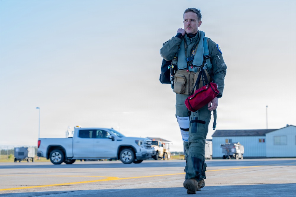 A military pilot in full flight gear walks across an airfield tarmac, carrying a red bag and adjusting his collar or communication equipment. He is dressed in an olive green flight suit with a tactical vest, survival gear, and what appears to be a G-suit leg attachment. The background shows ground support vehicles, airport buildings, and open sky consistent with a military air base environment.