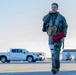 A military pilot in full flight gear walks across an airfield tarmac, carrying a red bag and adjusting his collar or communication equipment. He is dressed in an olive green flight suit with a tactical vest, survival gear, and what appears to be a G-suit leg attachment. The background shows ground support vehicles, airport buildings, and open sky consistent with a military air base environment.