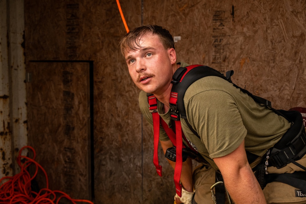 A young male wearing a green t-shirt and red safety harness is photographed in what appears to be a training or industrial environment with plywood walls. He appears physically exerted, with sweat visible on his face and hair, and is leaning forward in an alert posture. The setting and equipment suggest a firefighter or rescue training exercise.