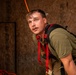 A young male wearing a green t-shirt and red safety harness is photographed in what appears to be a training or industrial environment with plywood walls. He appears physically exerted, with sweat visible on his face and hair, and is leaning forward in an alert posture. The setting and equipment suggest a firefighter or rescue training exercise.