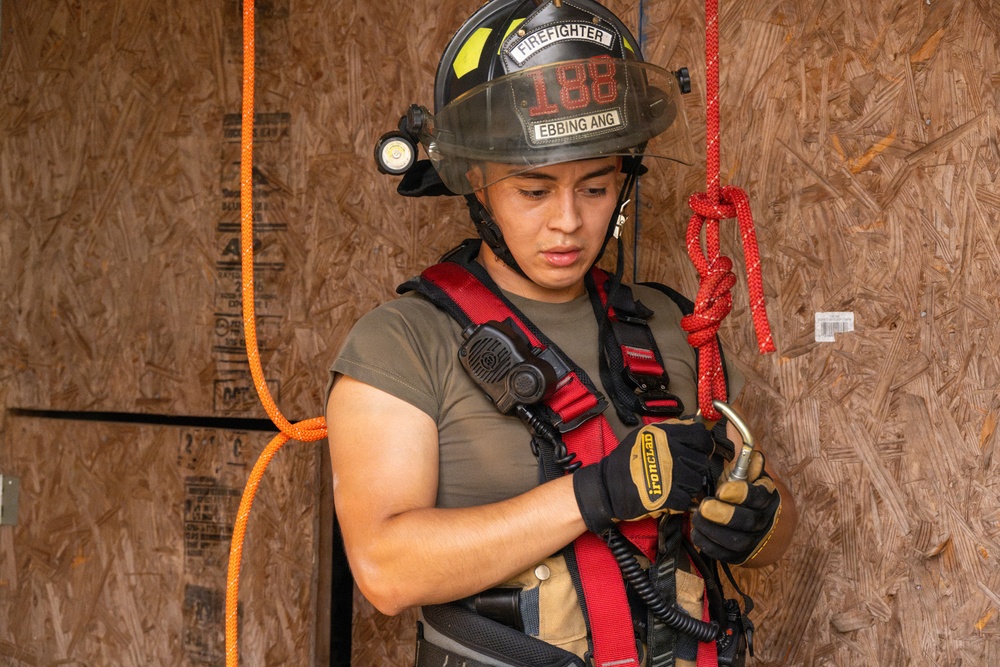 A firefighter wearing a black helmet labeled 'FIREFIGHTER 188' and 'EBBING ANG' is focused on connecting or checking a carabiner attached to a red safety harness. The individual is wearing Ironclad gloves and has a radio clipped to their chest. The setting appears to be a training structure made of oriented strand board (OSB).