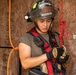 A firefighter wearing a black helmet labeled 'FIREFIGHTER 188' and 'EBBING ANG' is focused on connecting or checking a carabiner attached to a red safety harness. The individual is wearing Ironclad gloves and has a radio clipped to their chest. The setting appears to be a training structure made of oriented strand board (OSB).