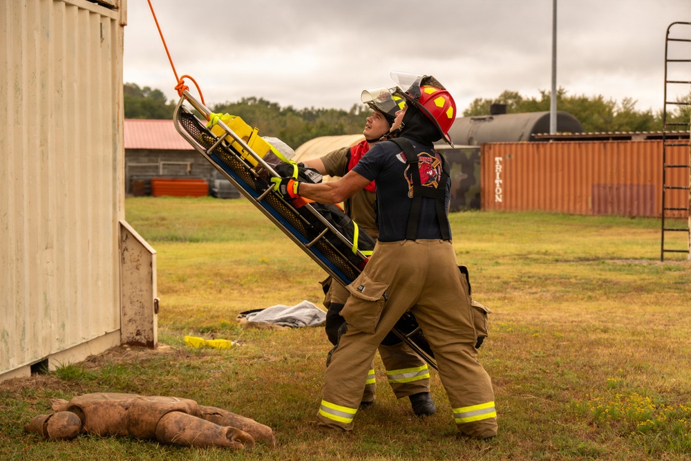 Two firefighters in full turnout gear are conducting a training exercise, handling a Stokes basket stretcher that is being lowered or raised via an orange rope attached to a structure. The scene takes place at an outdoor training facility with shipping containers, a tank, and various training props visible in the background. This appears to be a rope rescue or confined space rescue drill.
