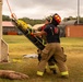 Two firefighters in full turnout gear are conducting a training exercise, handling a Stokes basket stretcher that is being lowered or raised via an orange rope attached to a structure. The scene takes place at an outdoor training facility with shipping containers, a tank, and various training props visible in the background. This appears to be a rope rescue or confined space rescue drill.