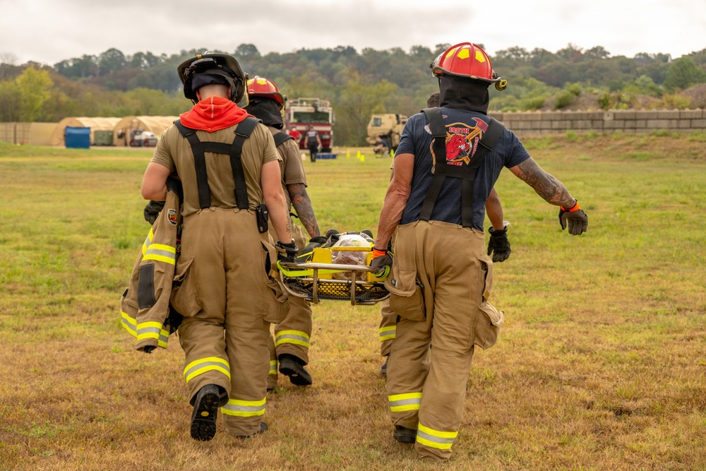 Three firefighters in full turnout gear are carrying a patient on a yellow stretcher across an open grassy field. The scene appears to be a mass casualty training exercise, evidenced by tents, military vehicles, and marked staging areas visible in the background. The responders are moving toward what appears to be a triage or treatment area.