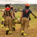 Three firefighters in full turnout gear are carrying a patient on a yellow stretcher across an open grassy field. The scene appears to be a mass casualty training exercise, evidenced by tents, military vehicles, and marked staging areas visible in the background. The responders are moving toward what appears to be a triage or treatment area.