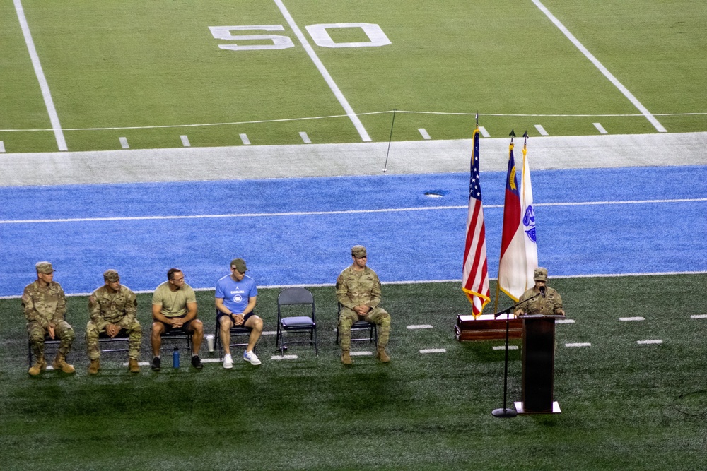 A military ceremony is taking place on the field of an American football stadium, near the 50-yard line. Several military personnel in camouflage uniforms are seated in folding chairs while one individual speaks at a podium flanked by flags. Two civilians in casual clothing are seated among the uniformed personnel, suggesting a mixed military-civilian event.
