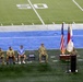 A military ceremony is taking place on the field of an American football stadium, near the 50-yard line. Several military personnel in camouflage uniforms are seated in folding chairs while one individual speaks at a podium flanked by flags. Two civilians in casual clothing are seated among the uniformed personnel, suggesting a mixed military-civilian event.