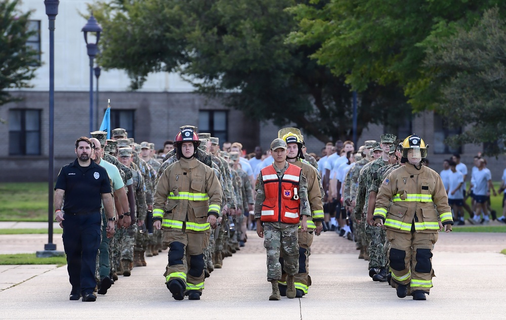 A large group of individuals in various uniforms are marching in formation along a paved walkway, likely during a commemorative or ceremonial event. The front row prominently features personnel in firefighter turnout gear, military camouflage, an EMS/medical vest, and a law enforcement uniform, suggesting a first responder tribute march. The setting appears to be a military installation or institutional campus with trees and brick buildings in the background.