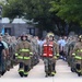 A large group of individuals in various uniforms are marching in formation along a paved walkway, likely during a commemorative or ceremonial event. The front row prominently features personnel in firefighter turnout gear, military camouflage, an EMS/medical vest, and a law enforcement uniform, suggesting a first responder tribute march. The setting appears to be a military installation or institutional campus with trees and brick buildings in the background.