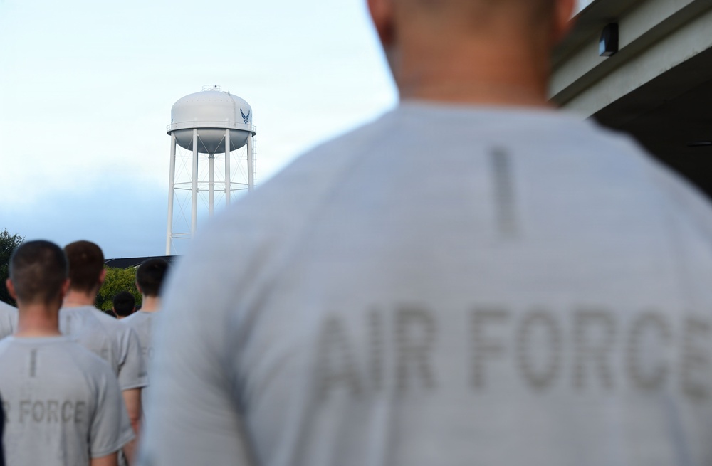 A group of individuals wearing gray 'Air Force' t-shirts are standing in formation, facing away from the camera. In the background, a white water tower bearing the United States Air Force logo (the Hap Arnold wing insignia) is prominently visible. The scene appears to take place on a U.S. Air Force military installation, likely during a physical training or formation activity.