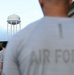 A group of individuals wearing gray 'Air Force' t-shirts are standing in formation, facing away from the camera. In the background, a white water tower bearing the United States Air Force logo (the Hap Arnold wing insignia) is prominently visible. The scene appears to take place on a U.S. Air Force military installation, likely during a physical training or formation activity.