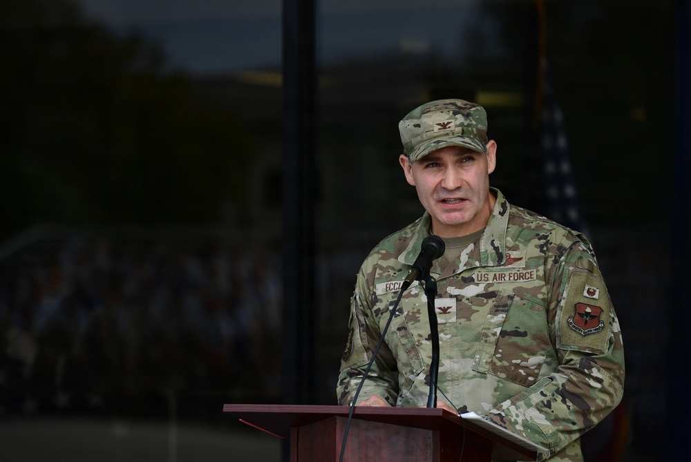 A U.S. Air Force officer in the rank of Colonel (identified by eagle insignia) is speaking at a podium with a microphone. The individual is wearing OCP (Operational Camouflage Pattern) uniform with 'U.S. AIR FORCE' tape and a name tape partially readable as 'ECCL-'. A formation of personnel is visible in the blurred background reflected in glass windows.