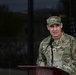 A U.S. Air Force officer in the rank of Colonel (identified by eagle insignia) is speaking at a podium with a microphone. The individual is wearing OCP (Operational Camouflage Pattern) uniform with 'U.S. AIR FORCE' tape and a name tape partially readable as 'ECCL-'. A formation of personnel is visible in the blurred background reflected in glass windows.