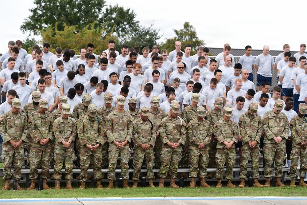 A large group of military personnel and trainees are assembled on bleachers in an outdoor setting, with heads bowed in what appears to be a moment of silence or prayer. The front rows consist of uniformed soldiers in camouflage (OCP/MultiCam) pattern uniforms and patrol caps, while the rows behind them wear white t-shirts, likely trainees or cadets. The setting suggests a formal military ceremony or memorial observance.