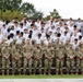 A large group of military personnel and trainees are assembled on bleachers in an outdoor setting, with heads bowed in what appears to be a moment of silence or prayer. The front rows consist of uniformed soldiers in camouflage (OCP/MultiCam) pattern uniforms and patrol caps, while the rows behind them wear white t-shirts, likely trainees or cadets. The setting suggests a formal military ceremony or memorial observance.