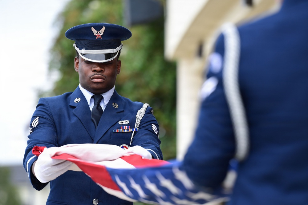 A United States Air Force honor guard member in dress blues is participating in a formal flag folding ceremony. The airman, wearing white gloves, is carefully folding an American flag with a second uniformed member visible in the foreground. The scene appears to be an official military ceremony, likely a funeral, memorial, or formal military event.