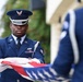 A United States Air Force honor guard member in dress blues is participating in a formal flag folding ceremony. The airman, wearing white gloves, is carefully folding an American flag with a second uniformed member visible in the foreground. The scene appears to be an official military ceremony, likely a funeral, memorial, or formal military event.