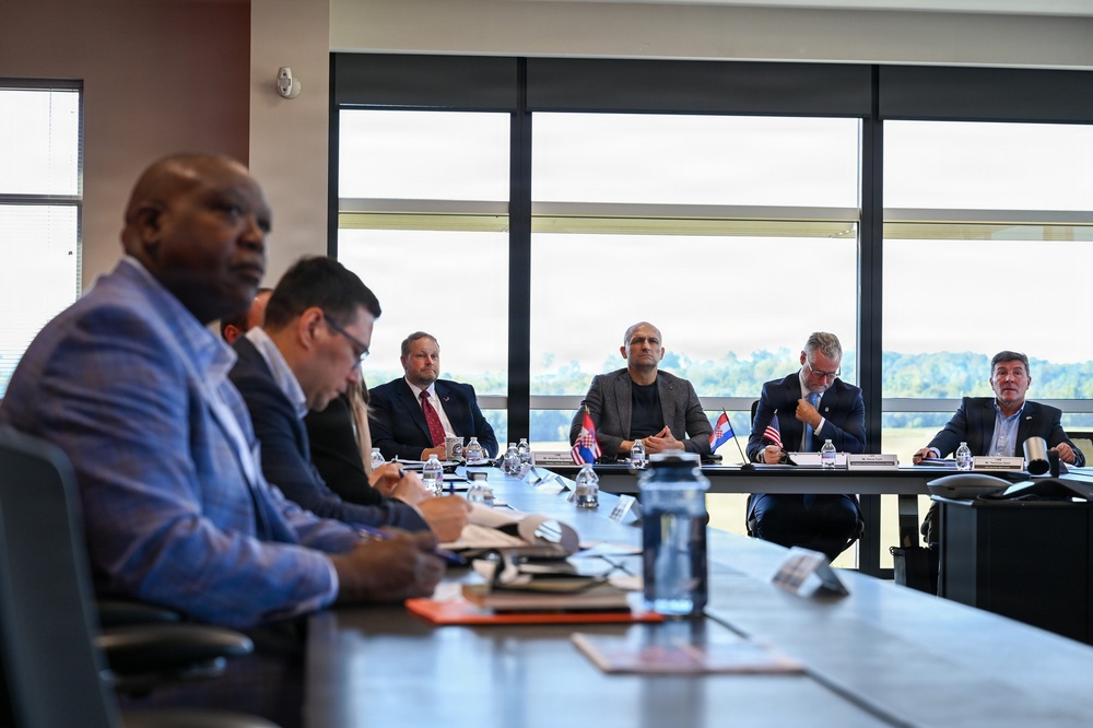 A formal meeting or delegation is taking place in a modern conference room with large windows overlooking a green landscape. Multiple individuals are seated around a long conference table, with small national flags visible — including what appear to be Croatian and American flags — suggesting an international or diplomatic meeting. Participants are engaged in note-taking, reviewing documents, and attentive listening.