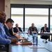 A formal meeting or delegation is taking place in a modern conference room with large windows overlooking a green landscape. Multiple individuals are seated around a long conference table, with small national flags visible — including what appear to be Croatian and American flags — suggesting an international or diplomatic meeting. Participants are engaged in note-taking, reviewing documents, and attentive listening.