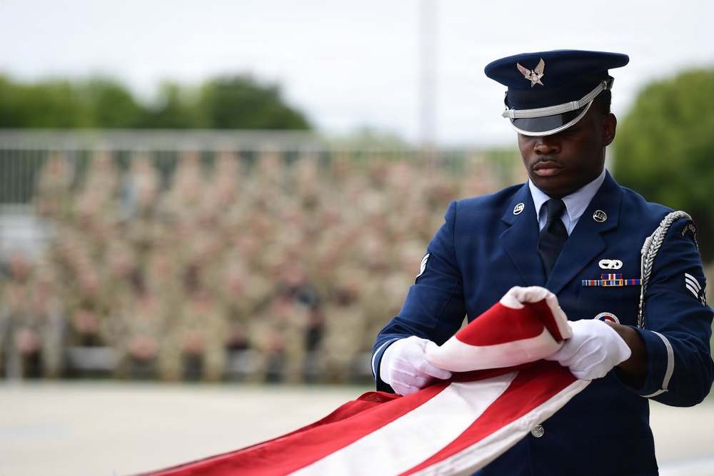 A United States Air Force service member in dress blues uniform is carefully folding an American flag during what appears to be a formal military ceremony. The airman is wearing white gloves and handling the flag with precision and solemnity. A large formation of uniformed personnel is visible in the blurred background, indicating a significant ceremonial event.