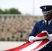 A United States Air Force service member in dress blues uniform is carefully folding an American flag during what appears to be a formal military ceremony. The airman is wearing white gloves and handling the flag with precision and solemnity. A large formation of uniformed personnel is visible in the blurred background, indicating a significant ceremonial event.