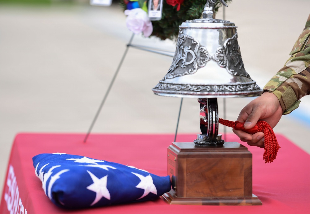 A military ceremony is taking place, featuring a soldier in camouflage uniform ringing an ornate silver bell mounted on a wooden base. A folded American flag rests on a red-draped table alongside the bell, indicating a formal memorial or honors ceremony. In the background, a wreath with photographs and flowers is visible on a stand.