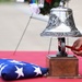 A military ceremony is taking place, featuring a soldier in camouflage uniform ringing an ornate silver bell mounted on a wooden base. A folded American flag rests on a red-draped table alongside the bell, indicating a formal memorial or honors ceremony. In the background, a wreath with photographs and flowers is visible on a stand.
