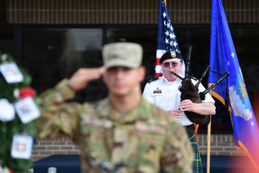 A military memorial or ceremonial event is taking place outdoors, featuring a soldier in camouflage uniform rendering a salute in the foreground and a bagpiper in white dress uniform with a green tartan kilt playing bagpipes in the background. An American flag and what appears to be a United States Air Force flag are visible alongside a memorial wreath with photographs. The name tag on the bagpiper's uniform reads 'CHASTANT'.