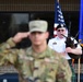 A military memorial or ceremonial event is taking place outdoors, featuring a soldier in camouflage uniform rendering a salute in the foreground and a bagpiper in white dress uniform with a green tartan kilt playing bagpipes in the background. An American flag and what appears to be a United States Air Force flag are visible alongside a memorial wreath with photographs. The name tag on the bagpiper's uniform reads 'CHASTANT'.