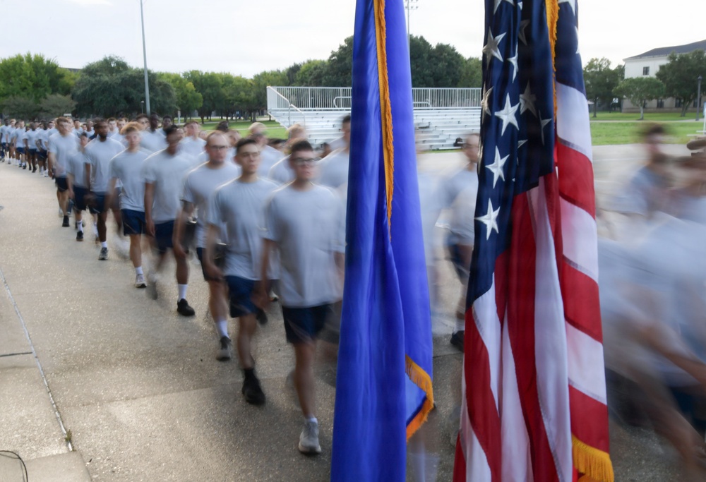 A large formation of military trainees or cadets in physical training uniforms (white t-shirts and blue shorts) marches in formation past two flags. The foreground prominently displays the United States flag and what appears to be the U.S. Air Force flag with gold fringe. The image uses motion blur to convey movement and scale of the formation.