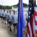 A large formation of military trainees or cadets in physical training uniforms (white t-shirts and blue shorts) marches in formation past two flags. The foreground prominently displays the United States flag and what appears to be the U.S. Air Force flag with gold fringe. The image uses motion blur to convey movement and scale of the formation.