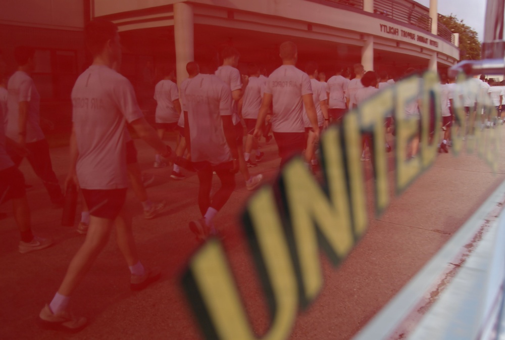 A group of people wearing matching white t-shirts are walking along a sidewalk in front of a building. The image is taken through a glass surface displaying the word 'UNITED' in yellow and black lettering, creating a reflection effect with a red tint overlay. The building signage in the background appears reversed, confirming the glass reflection perspective.