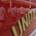 A group of people wearing matching white t-shirts are walking along a sidewalk in front of a building. The image is taken through a glass surface displaying the word 'UNITED' in yellow and black lettering, creating a reflection effect with a red tint overlay. The building signage in the background appears reversed, confirming the glass reflection perspective.