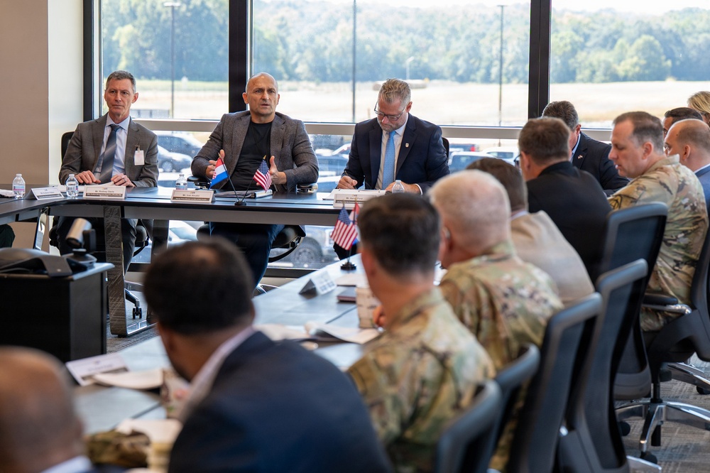 A formal bilateral or multinational military and civilian meeting is taking place in a modern conference room. Small flags including the United States flag and what appears to be a Luxembourg or Netherlands flag are visible on the table, suggesting an international defense or diplomatic engagement. A mix of uniformed military personnel in camouflage and civilian officials in suits are seated around a large conference table.