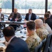 A formal bilateral or multinational military and civilian meeting is taking place in a modern conference room. Small flags including the United States flag and what appears to be a Luxembourg or Netherlands flag are visible on the table, suggesting an international defense or diplomatic engagement. A mix of uniformed military personnel in camouflage and civilian officials in suits are seated around a large conference table.