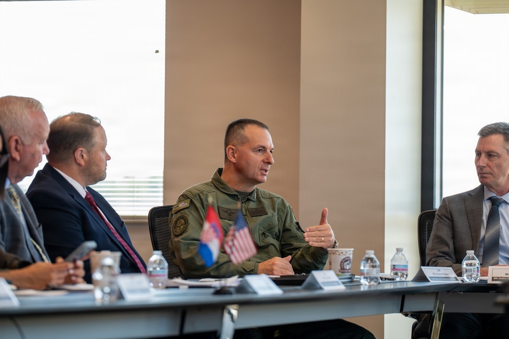 A formal meeting or briefing is taking place at a conference table, featuring a military officer in olive green flight suit as the apparent speaker. Small desktop flags representing the United States and what appears to be Serbia are visible on the table, suggesting a bilateral or international military/diplomatic engagement. Several civilians in business attire are seated alongside the uniformed officer.