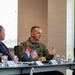 A formal meeting or briefing is taking place at a conference table, featuring a military officer in olive green flight suit as the apparent speaker. Small desktop flags representing the United States and what appears to be Serbia are visible on the table, suggesting a bilateral or international military/diplomatic engagement. Several civilians in business attire are seated alongside the uniformed officer.