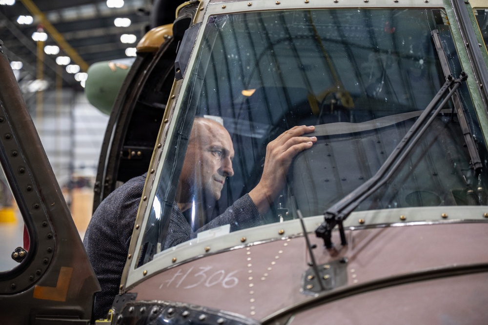 A man is inspecting or working on the interior of an aircraft cockpit from inside, visible through the curved glass canopy. The aircraft appears to be a vintage or historical military aircraft undergoing restoration or maintenance in a large hangar facility. The fuselage bears the marking 'H306' and shows riveted metal construction typical of mid-20th century aircraft.