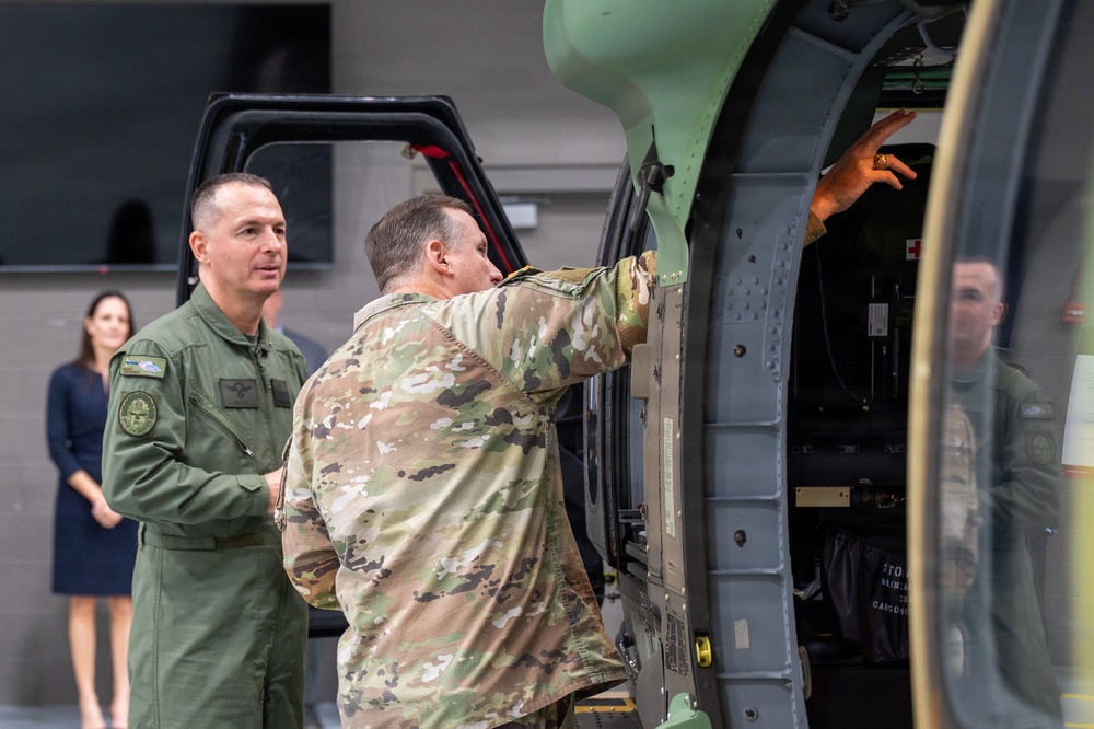 Two military personnel are examining the interior of a military helicopter, likely during an inspection or demonstration in an indoor hangar facility. One individual is wearing an olive green flight suit with patches, while the other is in multicam/OCP camouflage uniform and is pointing into the aircraft's open compartment. A civilian woman in a dark dress is visible in the background observing the interaction.