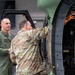 Two military personnel are examining the interior of a military helicopter, likely during an inspection or demonstration in an indoor hangar facility. One individual is wearing an olive green flight suit with patches, while the other is in multicam/OCP camouflage uniform and is pointing into the aircraft's open compartment. A civilian woman in a dark dress is visible in the background observing the interaction.