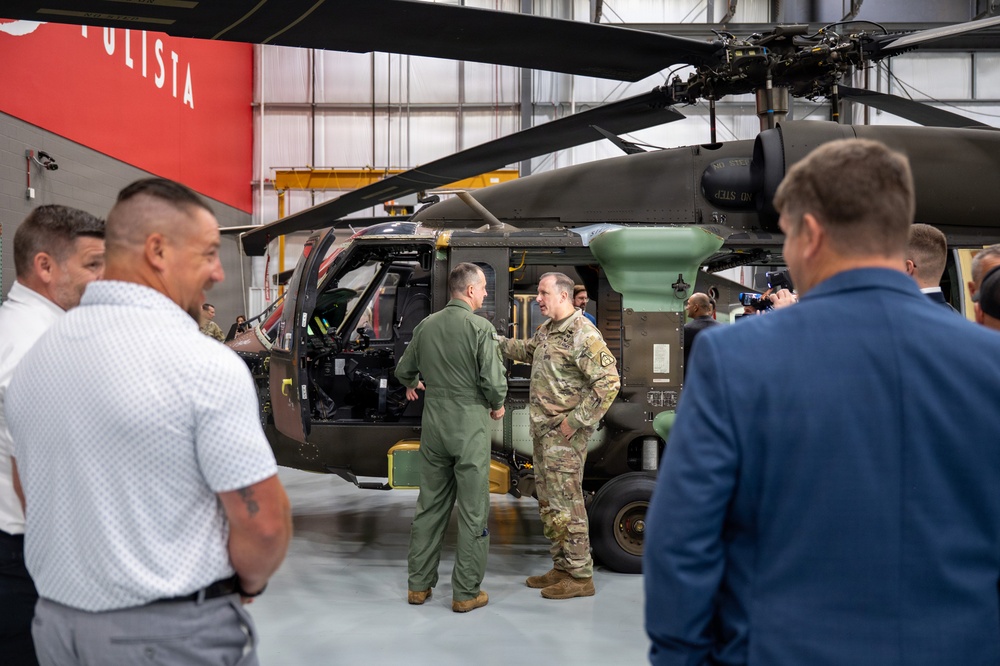 A military and civilian delegation is gathered inside a hangar facility, examining a military helicopter, likely a Sikorsky UH-60 Black Hawk. Two military personnel — one in a green flight suit and one in Army combat uniform (ACU/OCP pattern) — are engaged in conversation near the open cockpit of the aircraft. A red banner partially visible in the background reads 'ULISTA,' likely part of the 'SIKORSKY' or 'AGUSTAWESTLAND' branding.