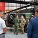 A military and civilian delegation is gathered inside a hangar facility, examining a military helicopter, likely a Sikorsky UH-60 Black Hawk. Two military personnel — one in a green flight suit and one in Army combat uniform (ACU/OCP pattern) — are engaged in conversation near the open cockpit of the aircraft. A red banner partially visible in the background reads 'ULISTA,' likely part of the 'SIKORSKY' or 'AGUSTAWESTLAND' branding.